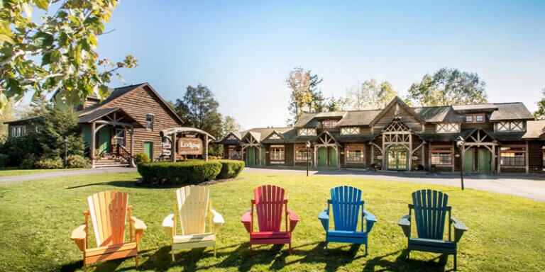 A row of five multicolored adirondack style chairs are seated neatly in front of the lawn of the Finger Lakes Lodge by Mirbeau in Skaneateles